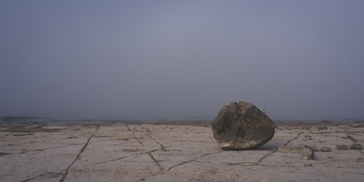 Photograph of a lone boulder resting on a wide rock shelf, framed in soft light. Suggests Sisyphus’s boulder, yet grounded on a level plane that hints the struggle may be achievable, not endless.