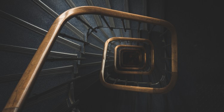 Photograph of a shadowed spiral staircase seen from above, wooden railing contrasting a blue-grey carpet, evoking descent and reflection.