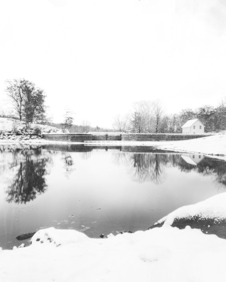 Long-exposure pinhole photograph looking downstream toward the Lockstation House at Jones Falls on the Rideau Canal, open water reflecting snowy shoreline and early winter sky.