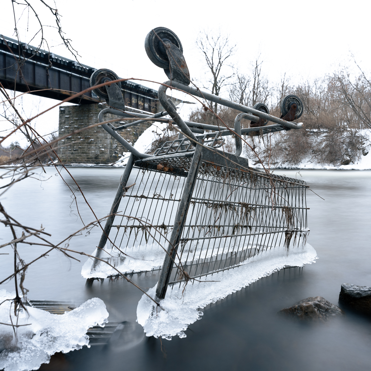 An overturned shopping cart frozen at the edge of a river in winter, partially embedded in ice, with smooth long exposure water surrounding it and a rail bridge visible in the background.