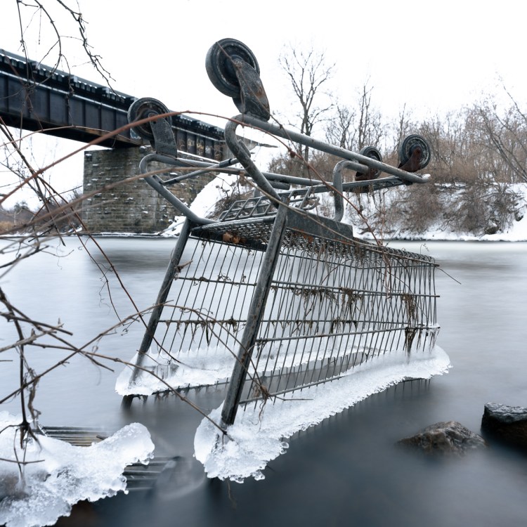 An overturned shopping cart frozen at the edge of a river in winter, partially embedded in ice, with smooth long exposure water surrounding it and a rail bridge visible in the background.