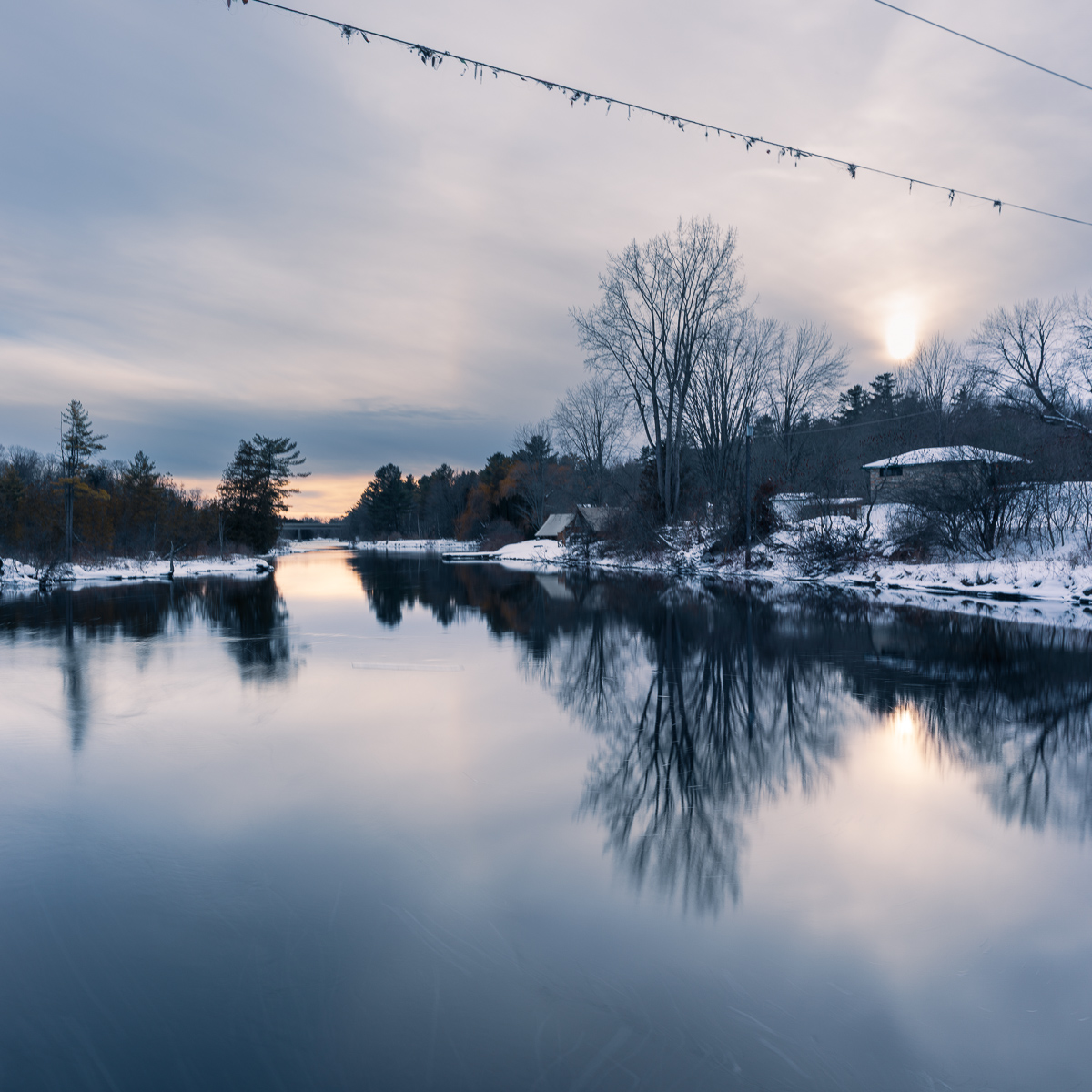 A winter river at dusk with snow along the banks and bare trees reflected in still, partially frozen water. The low sun glows through thin cloud, and the sky remains light at 4:30 pm.