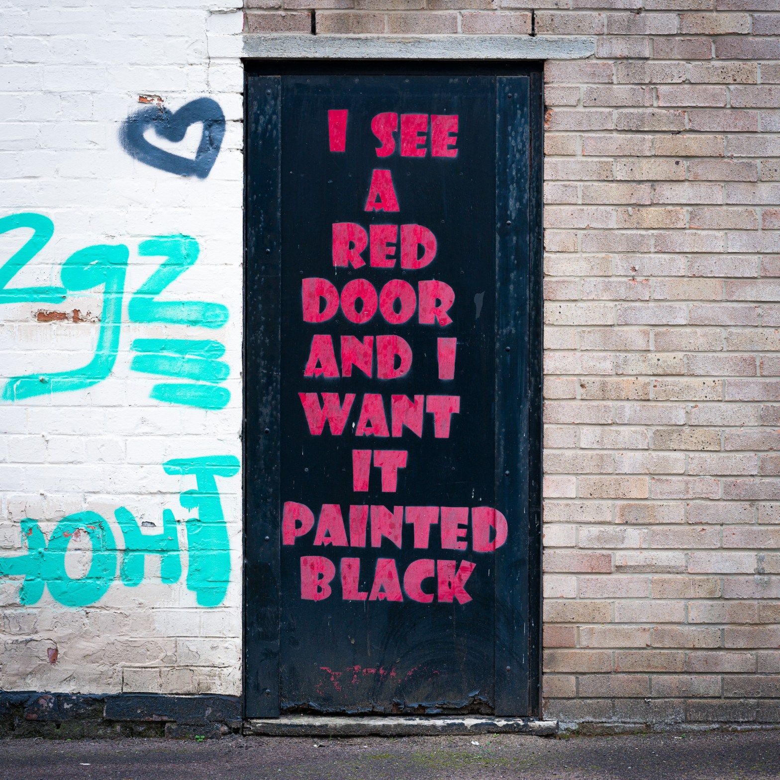 A black door in Bristol, UK, painted with pink text reading “I see a red door and I want it painted black,” set between white-painted brick with turquoise graffiti and pale brickwork.