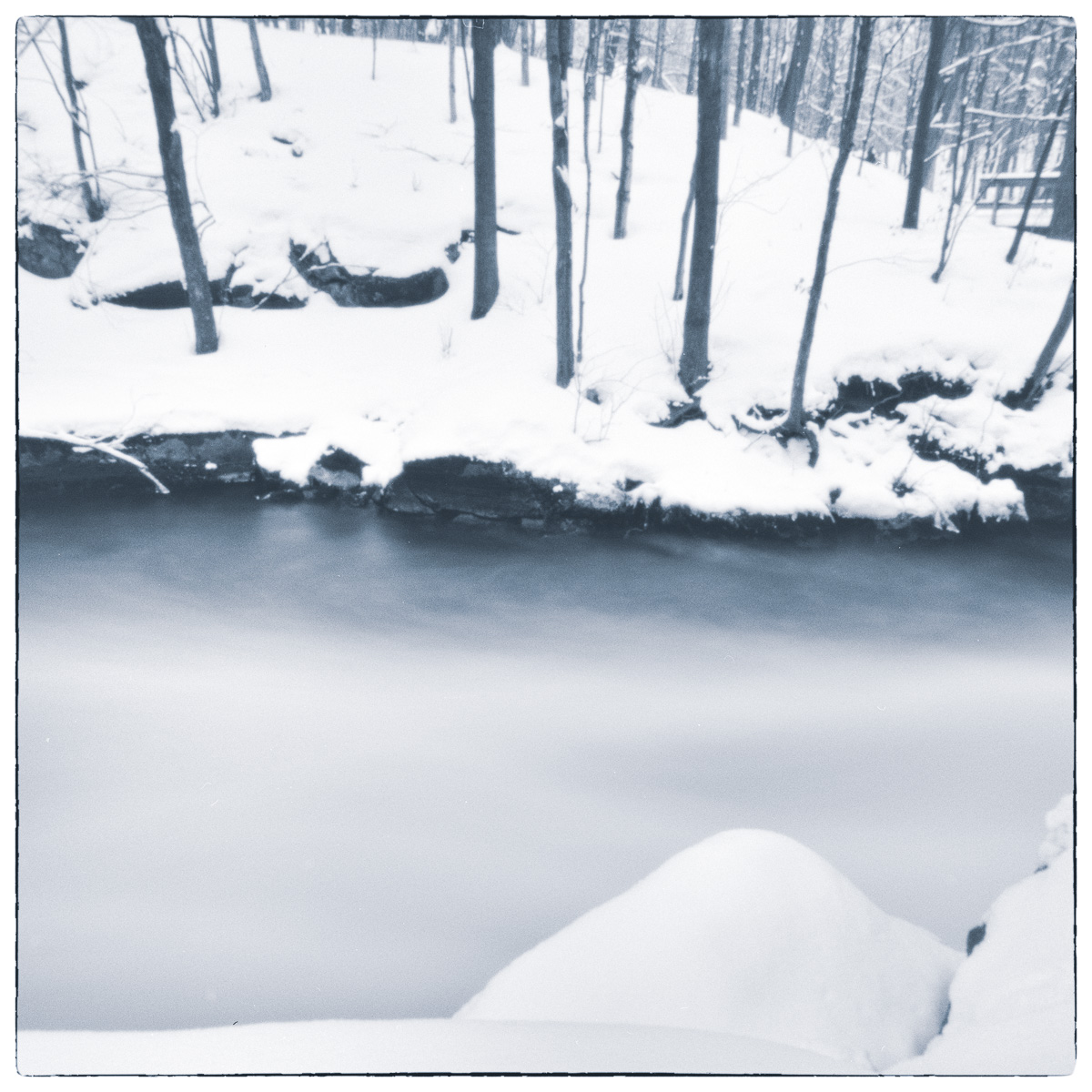 Snow-covered riverbank and blurred winter water beneath bare trees.