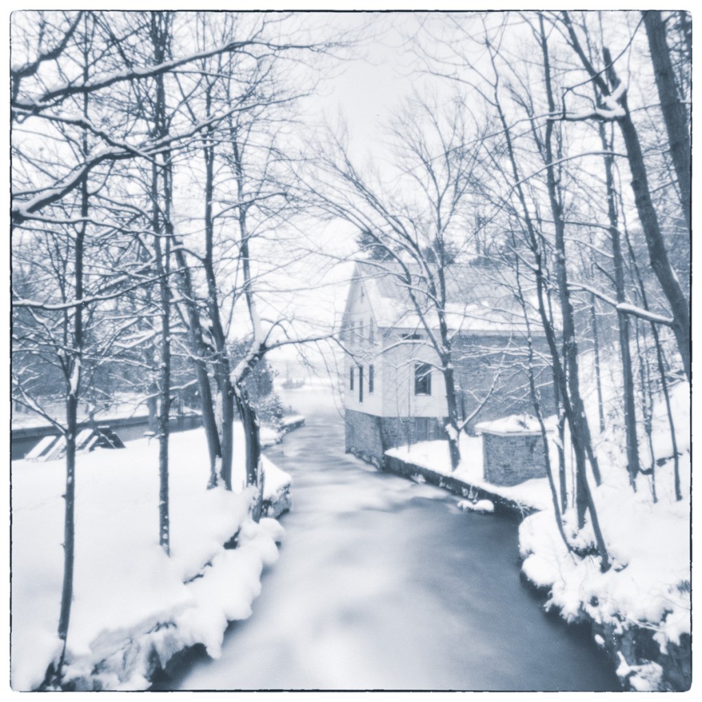 Snow-covered lockstation building and flowing winter water framed by bare trees.