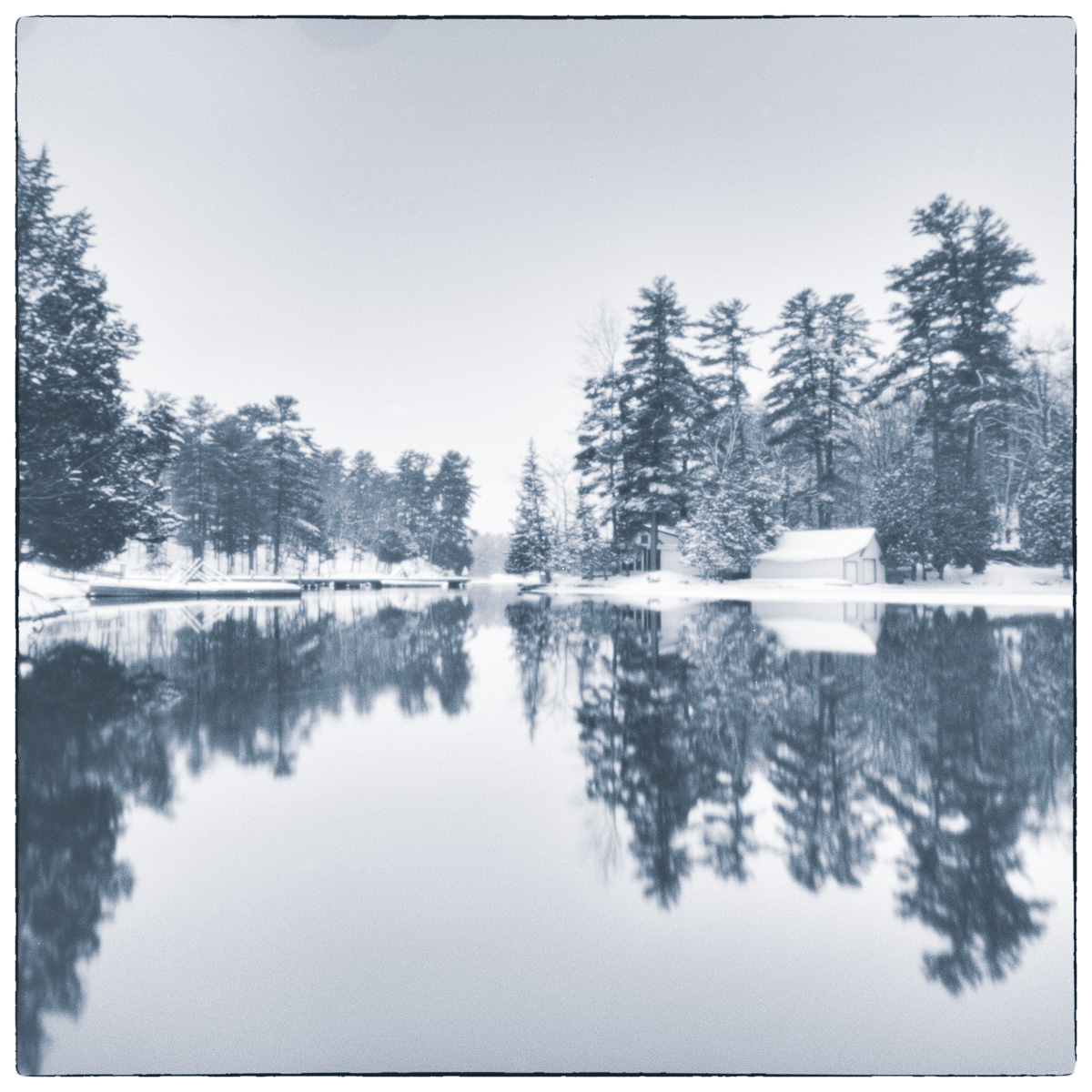 Winter canal landscape with snow-covered trees and buildings reflected in still water.