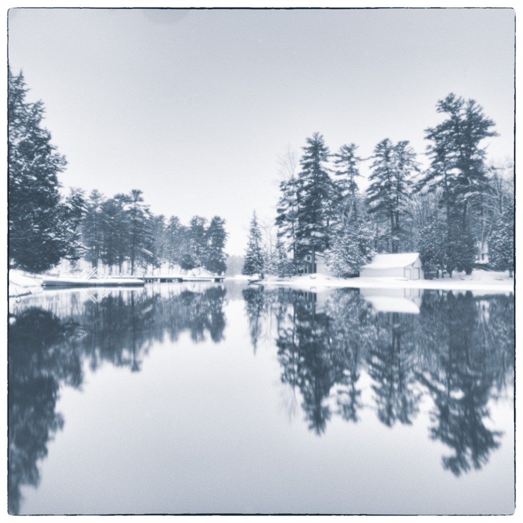 Winter canal landscape with snow-covered trees and buildings reflected in still water.