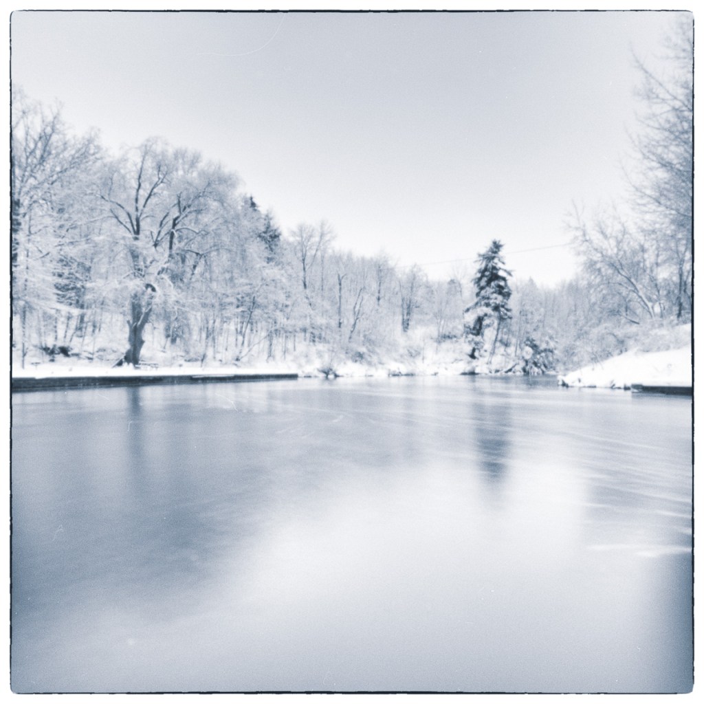 Winter river landscape with snow-covered trees and still water rendered in soft monochrome tones.