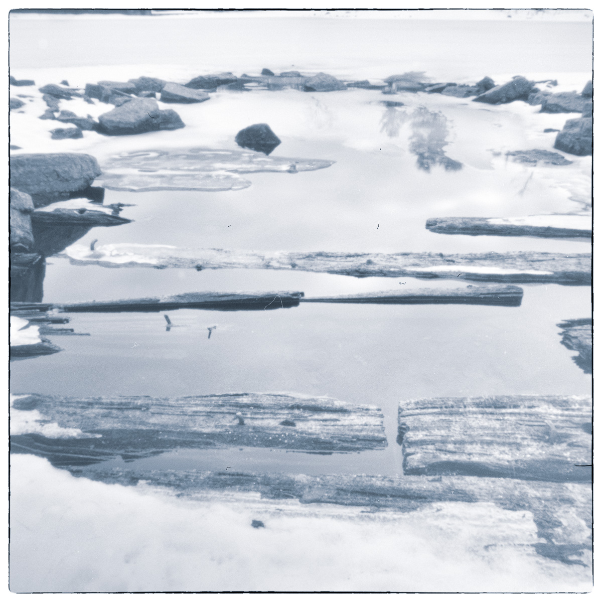 Long-exposure black and white photograph of meltwater, exposed wooden beams, rocks, and ice at the Jones Falls Lockstation basin.