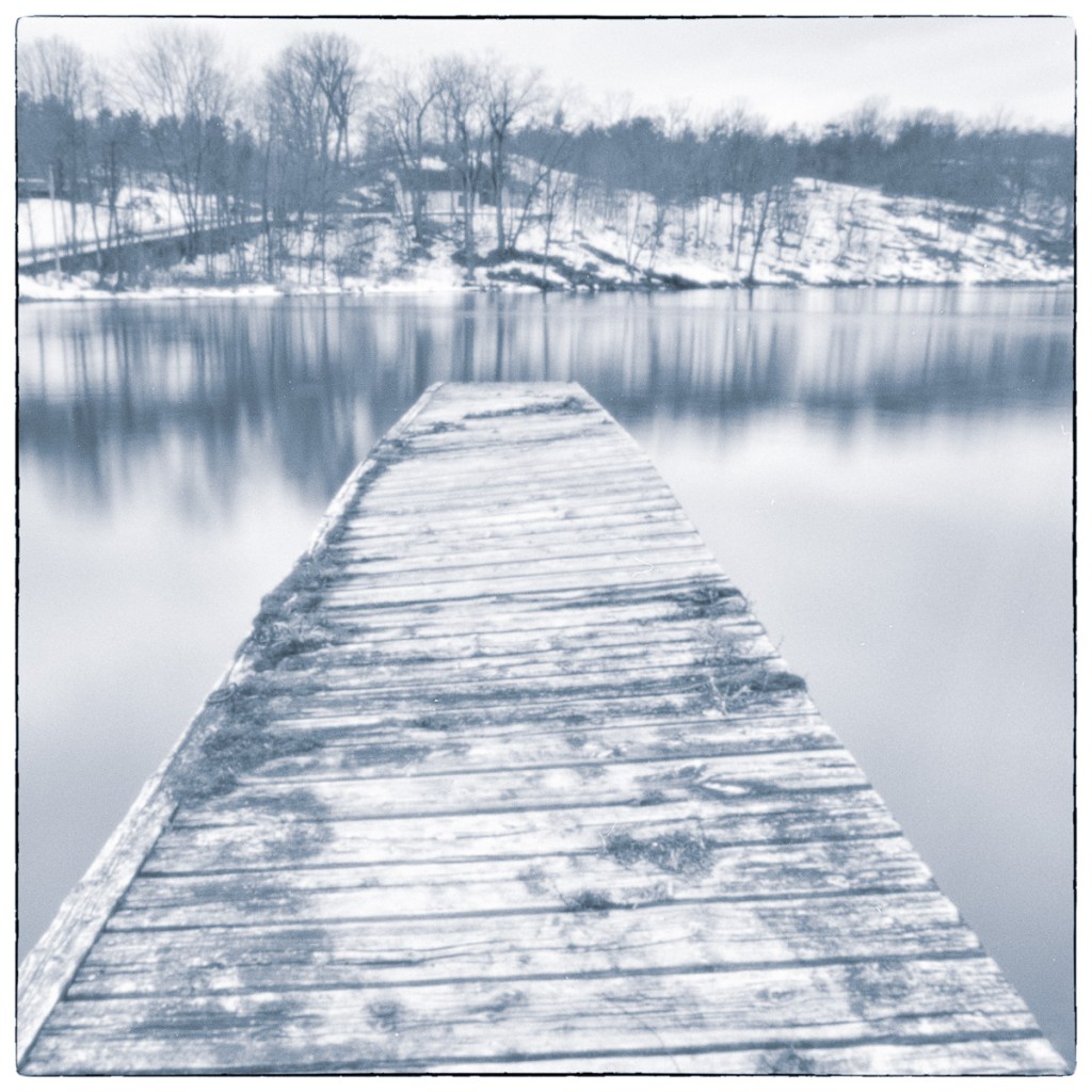Long-exposure black and white photograph of a weathered wooden dock extending into calm winter water at Jones Falls Bay, Ontario.