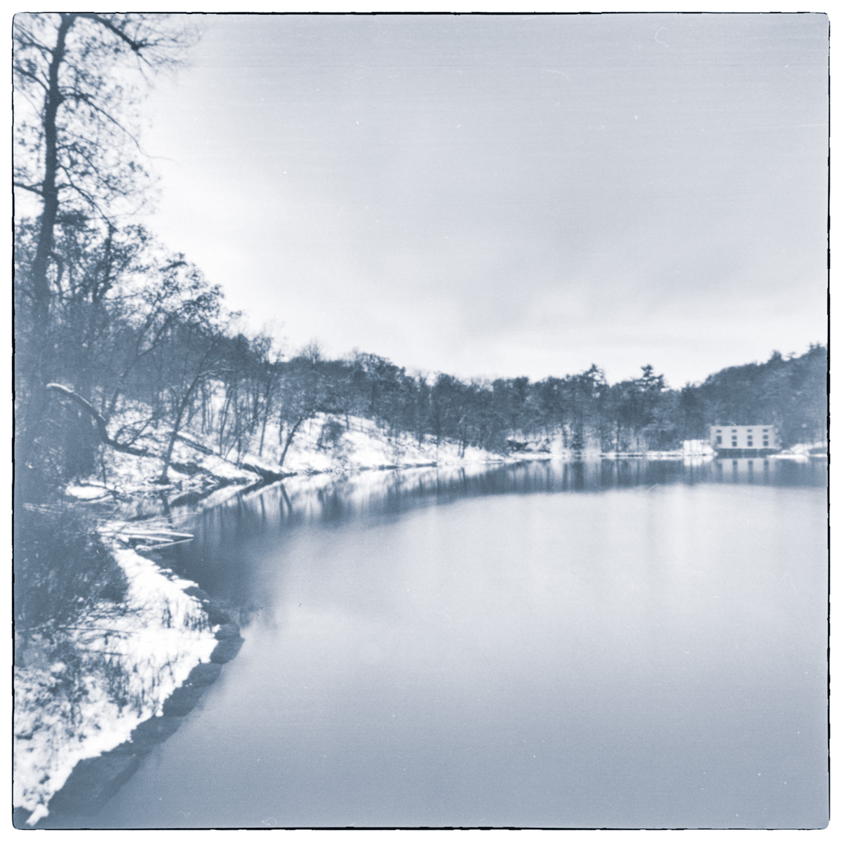 Long-exposure black and white photograph of the snow-covered shoreline at Jones Falls Bay during the season’s first snowfall, with calm water and a distant white building reflected across the bay.