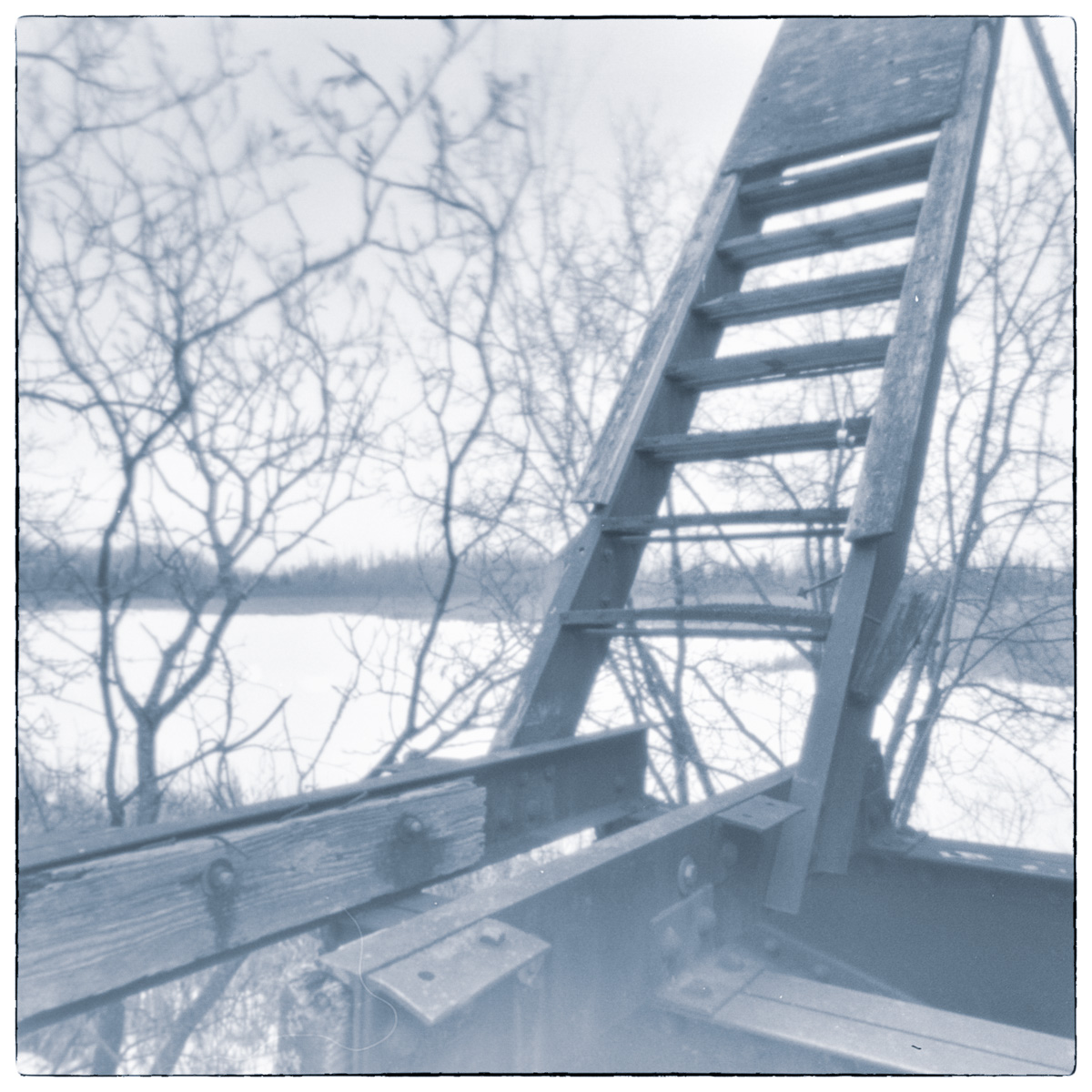 Long-exposure black and white photograph of the raised structure of the Smiths Falls Bascule Bridge, framed against bare winter trees and frozen water.