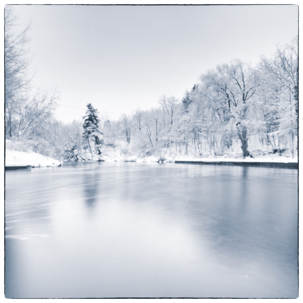 Long-exposure black and white photograph of calm winter water and snow-covered trees at Lower Brewers Lock, horizontally mirrored from a companion image in the sequence.