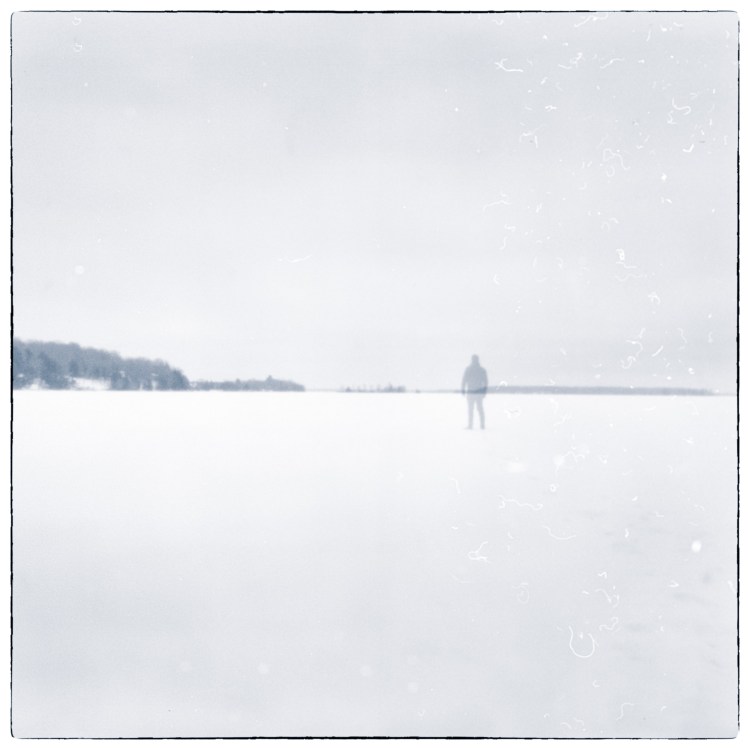 Solitary figure standing on a frozen landscape in winter, photographed in soft monochrome tones.