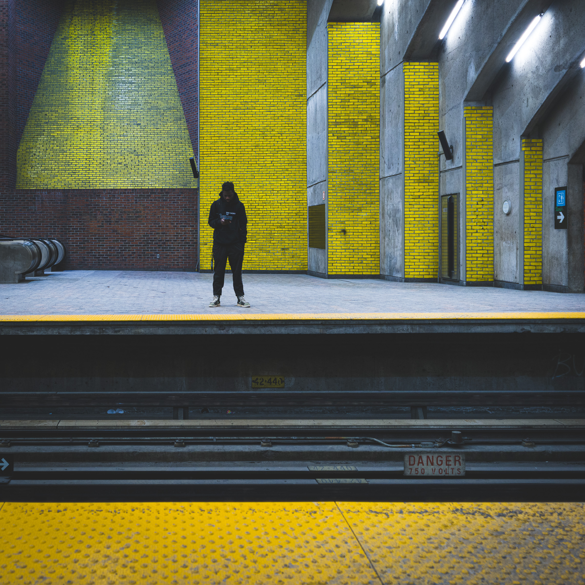 Colour photograph of a lone figure standing on the Saint-Henri platform in the Montréal Métro, surrounded by yellow brick walls and viewed from across the tracks.