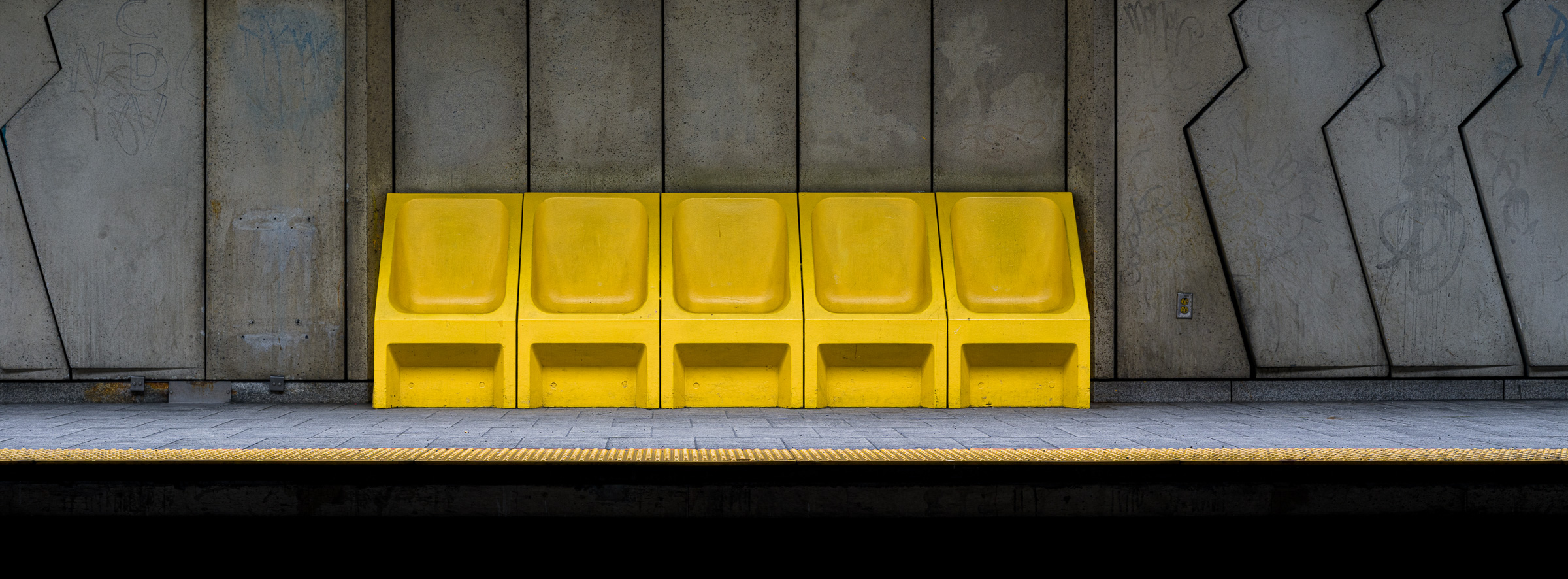Panoramic colour photograph of yellow platform seating against brutalist concrete walls at Côte-Sainte-Catherine station in the Montréal Métro, viewed from across the tracks.