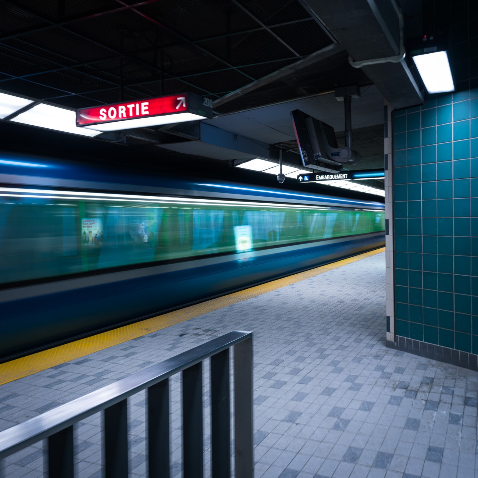 Long-exposure colour photograph of a train blurring past the Champ-de-Mars platform in the Montréal Métro, viewed from the platform entrance at the bottom of the stairs.