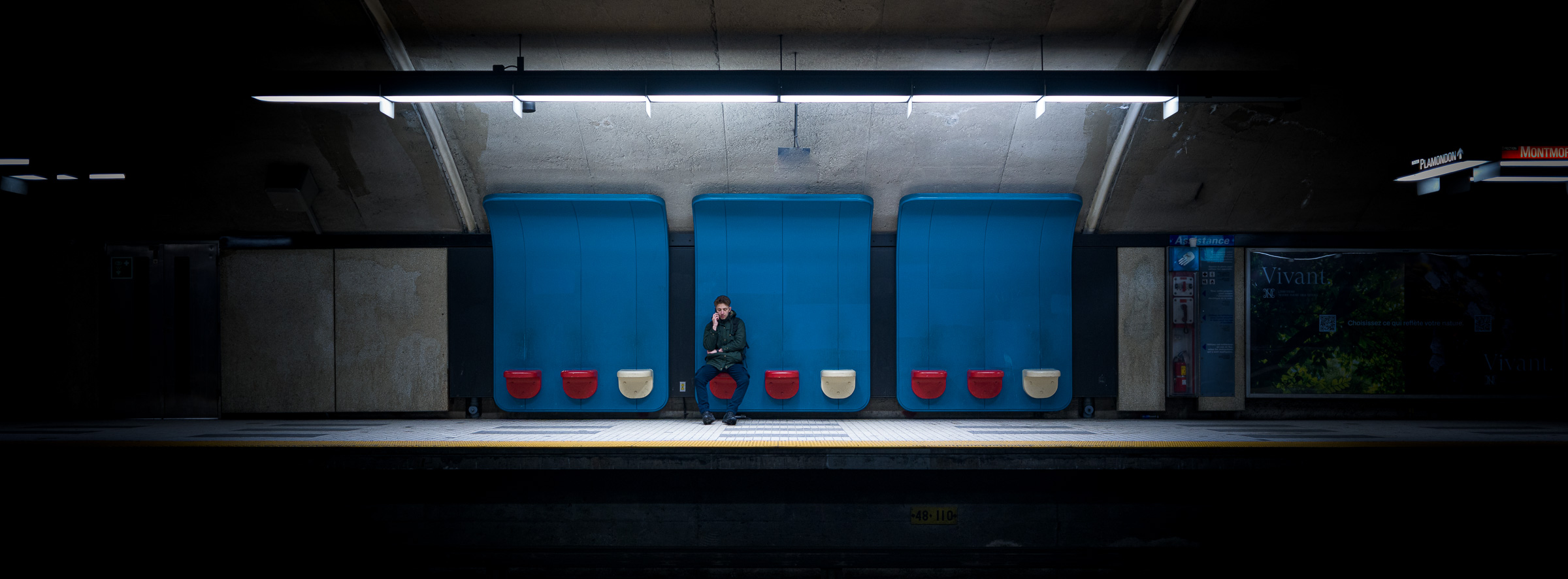 Panoramic colour photograph of a lone figure using a phone on the platform seating at Plamondon station in the Montréal Métro.