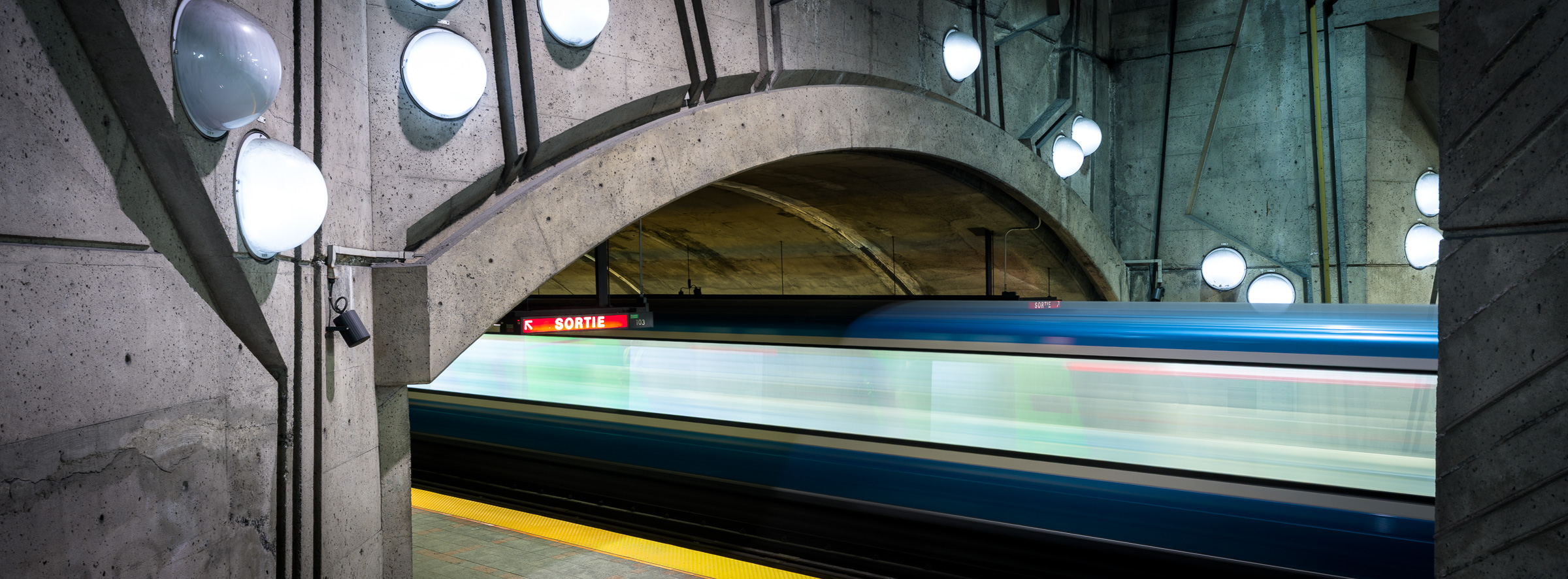 Panoramic long-exposure colour photograph of a train entering a brutalist concrete tunnel at De la Savane station in the Montréal Métro, with round dome lights on the walls.