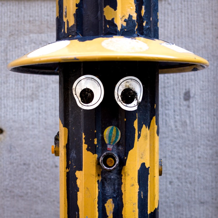 Googly eyes attached to a black and yellow post against a grey urban background.