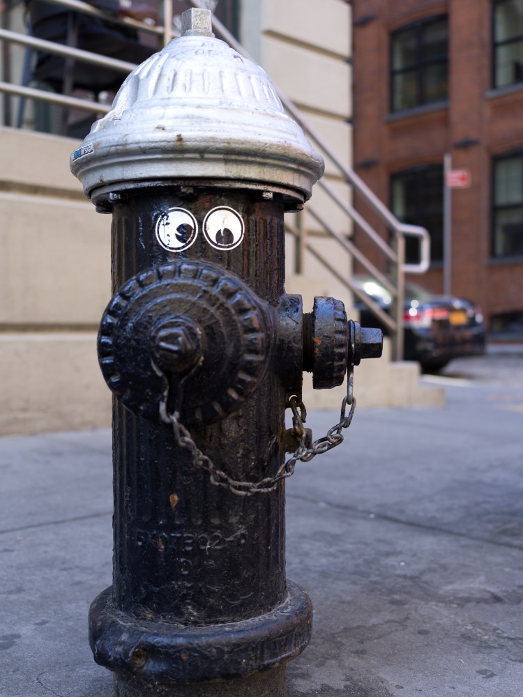 Black fire hydrant with googly eyes attached, photographed on a city sidewalk.