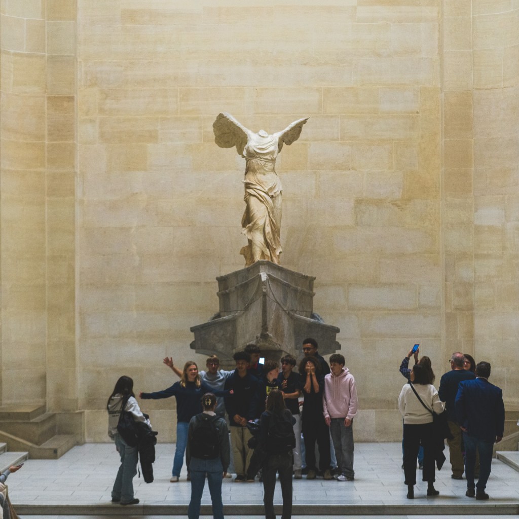 A handheld photograph of the Winged Victory of Samothrace at the Louvre, with visitors gathered at the base of the sculpture and several people raising phones to take pictures.