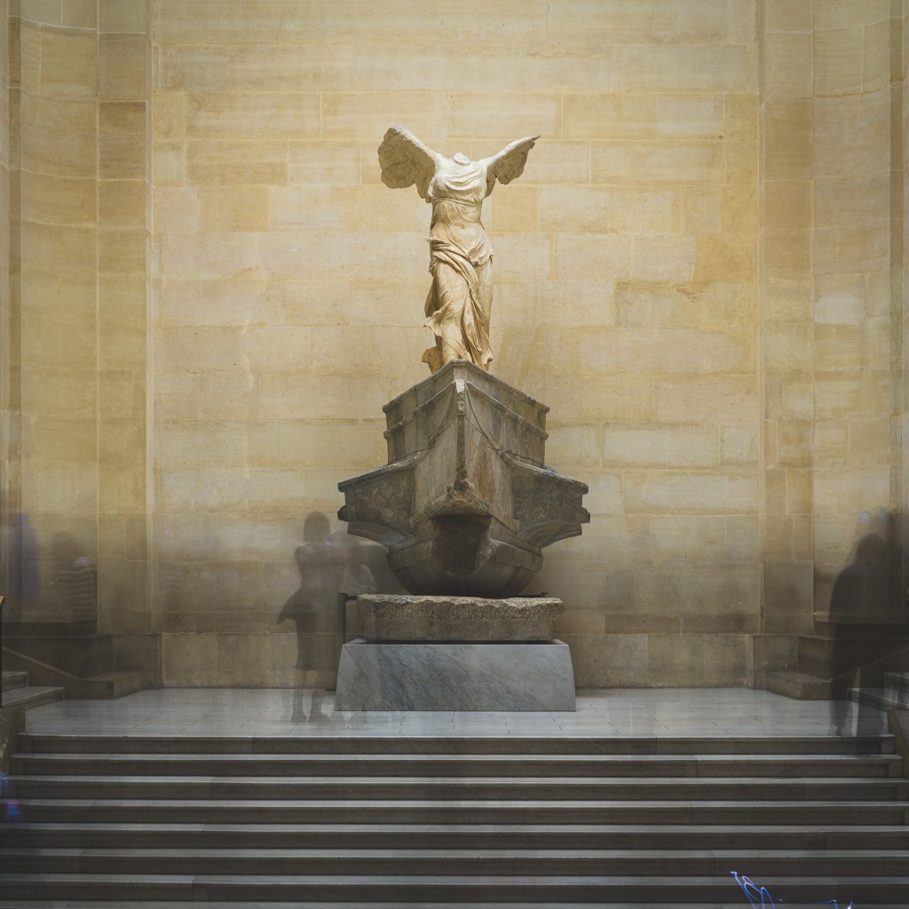 A 161-second long exposure of the Winged Victory of Samothrace at the Louvre, with visitors partially blurred into ghost-like figures around the sculpture.