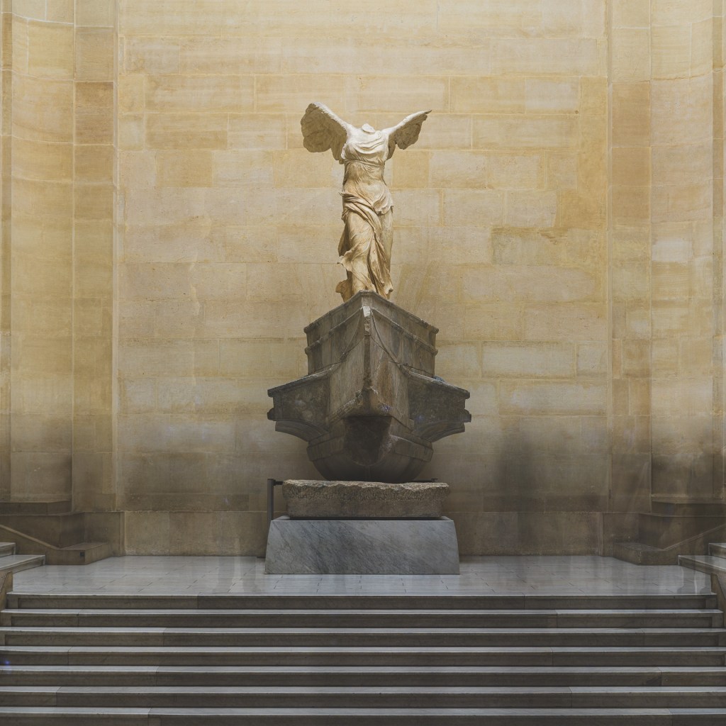 A 323-second long exposure of the Winged Victory of Samothrace at the Louvre, with the surrounding crowd dissolved into faint atmospheric traces.