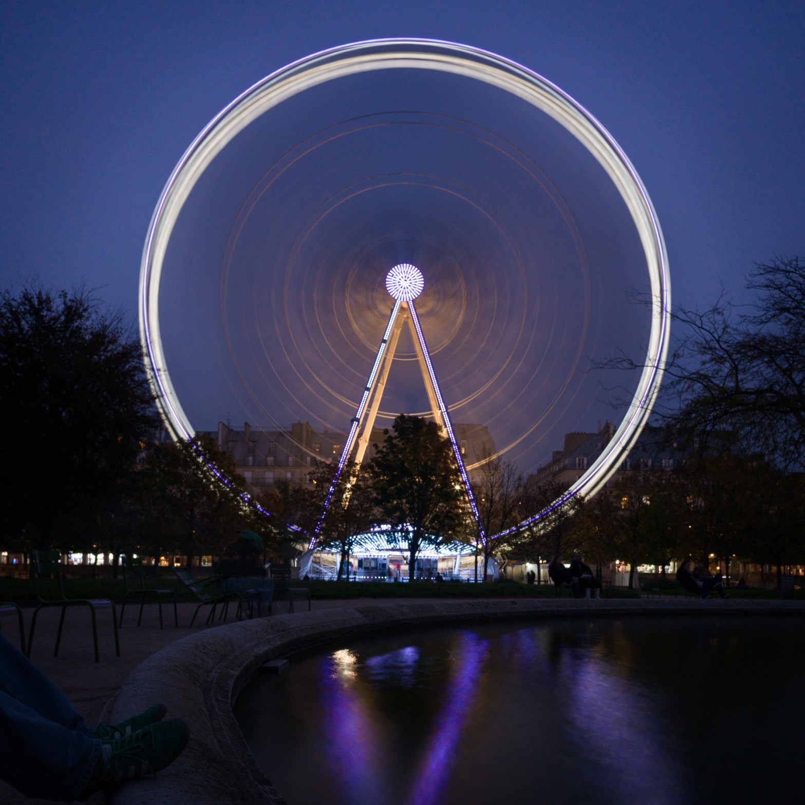 Long-exposure photograph of the Roue de Paris at dusk, with the Ferris wheel turning and its lights forming circular trails.