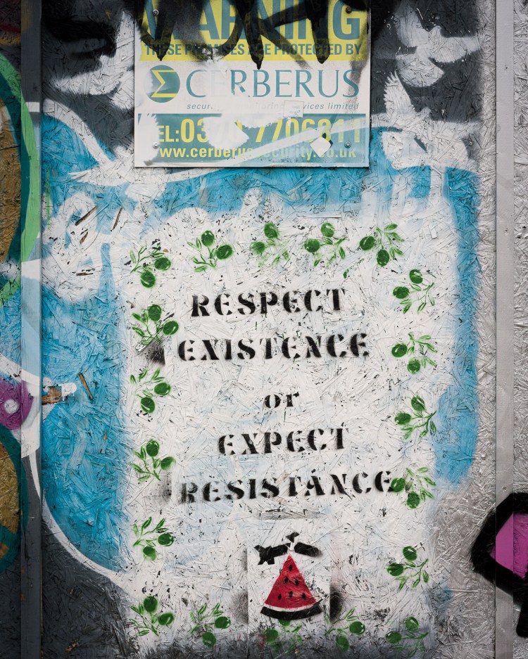 Stencil reading “Respect Existence or Expect Resistance” beneath security and warning signs.