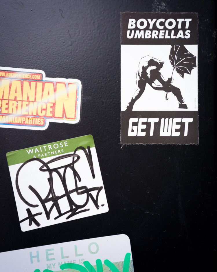 Sticker reading “Boycott Umbrellas / Get Wet” among layers of stickers on a Bristol street surface.