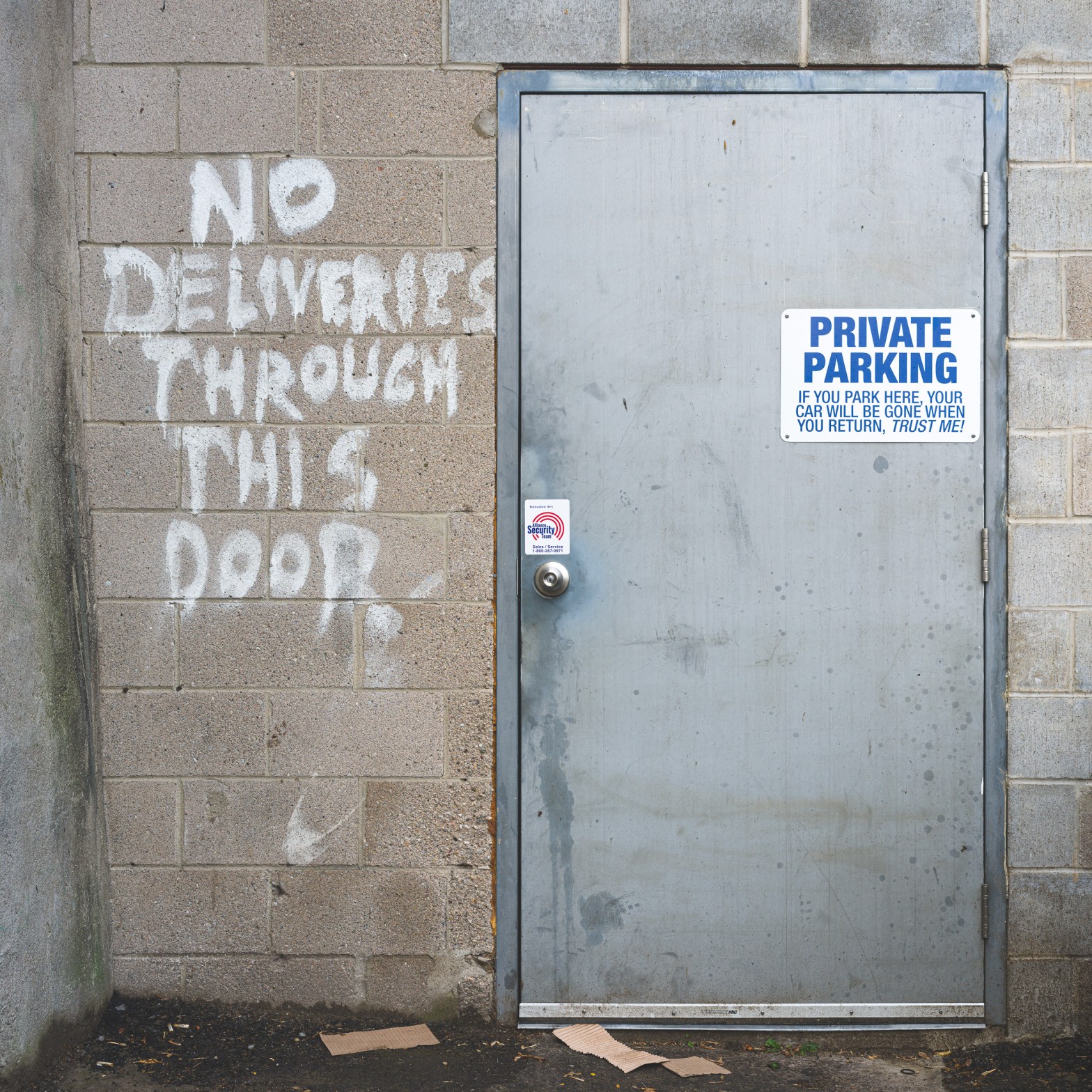 Grey metal service door in a Kingston alley beside hand-painted words reading “NO DELIVERIES THROUGH THIS DOOR.”