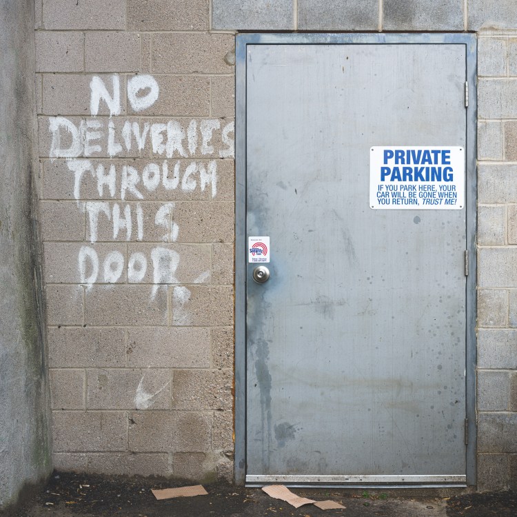 Grey metal service door in a Kingston alley beside hand-painted words reading “NO DELIVERIES THROUGH THIS DOOR.”