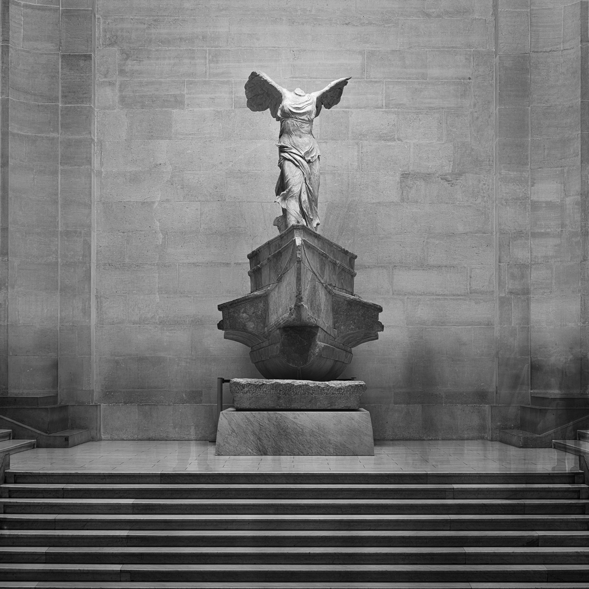 Long-exposure photograph of the Winged Victory of Samothrace at the Louvre, with passing visitors appearing as faint ghosted figures.