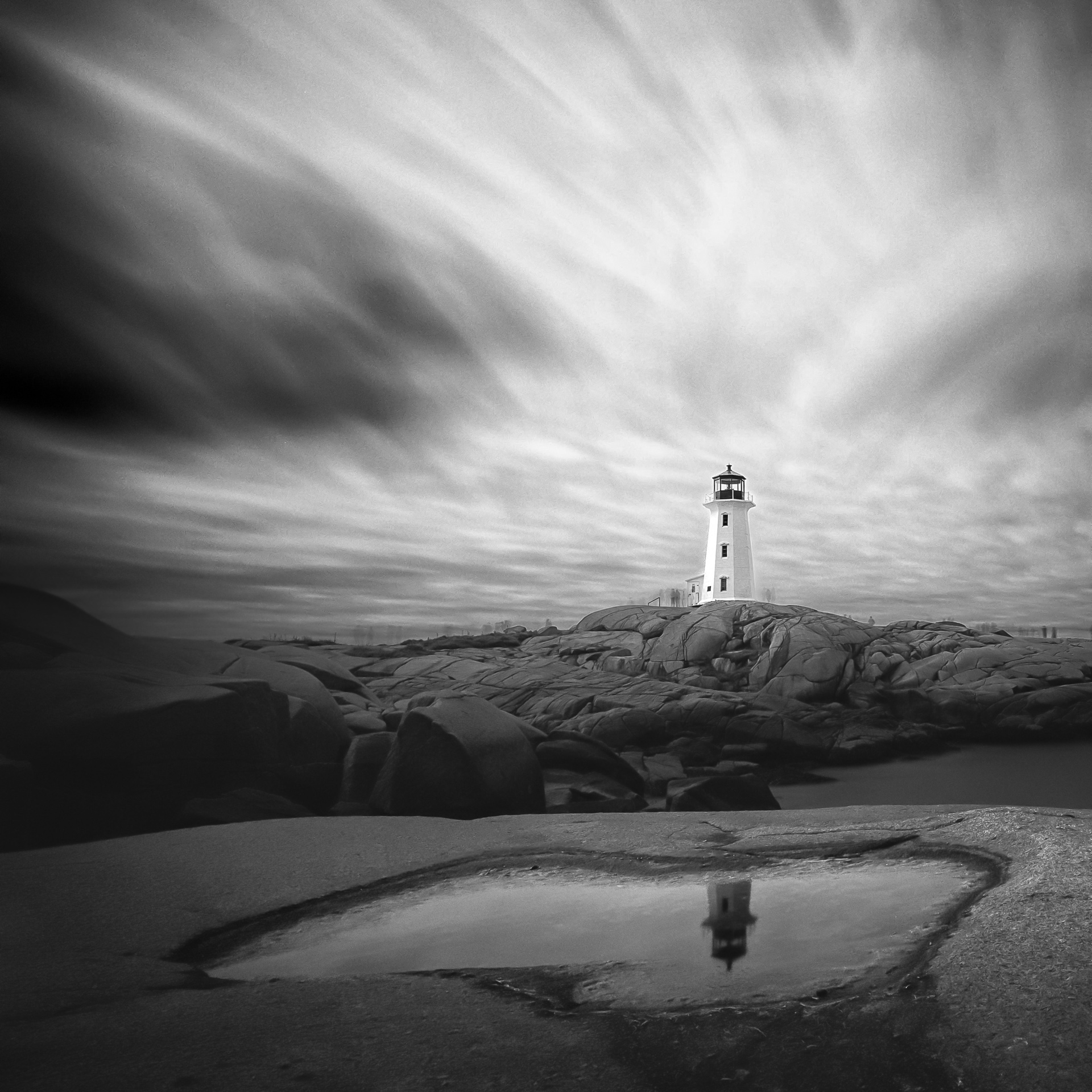 Black-and-white long exposure of Peggy’s Point Lighthouse at Peggy’s Cove, with streaked clouds and the lighthouse reflected in a foreground pool.