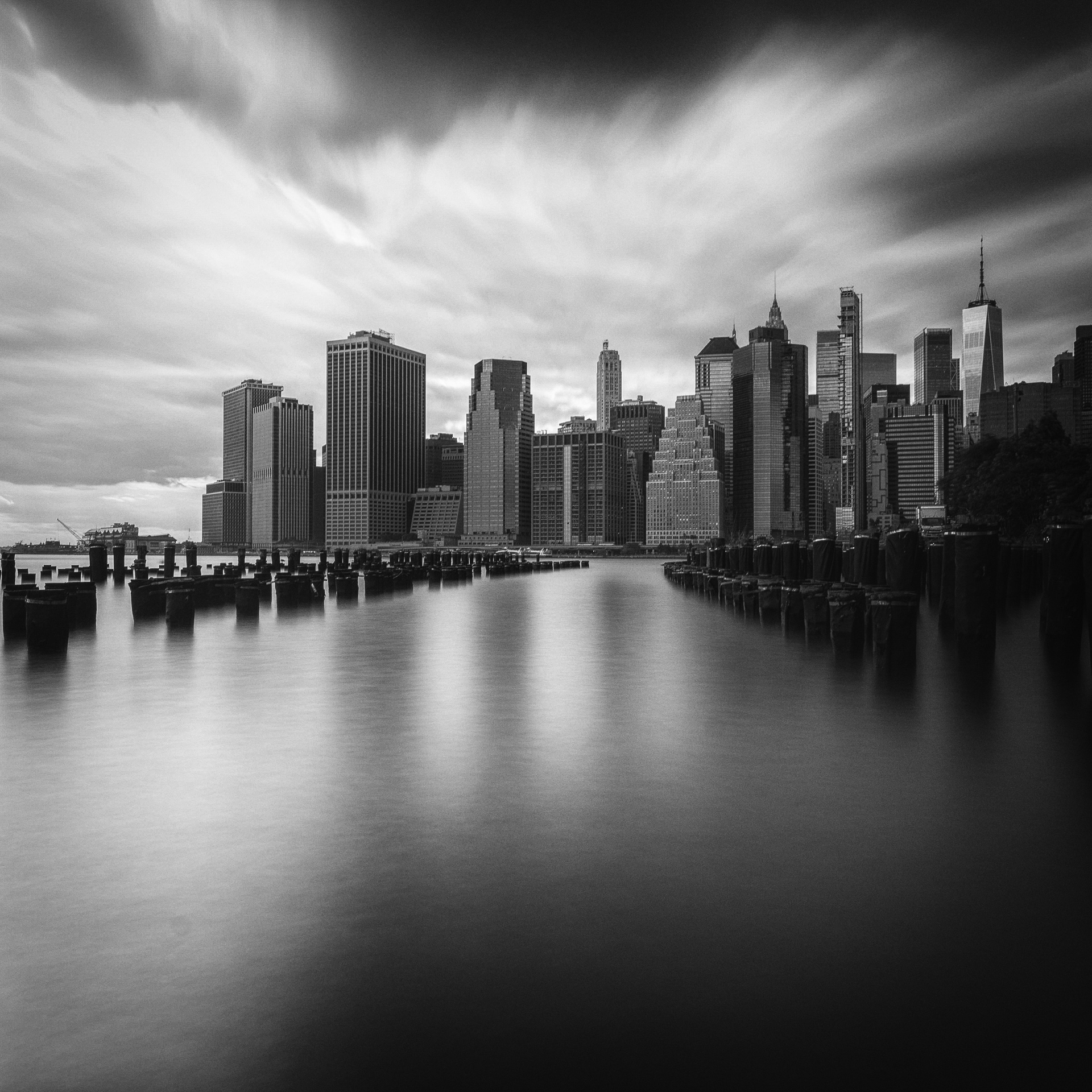 Black-and-white long exposure of the Lower Manhattan skyline seen across the East River, with smooth water and streaked clouds emphasizing the passage of time.