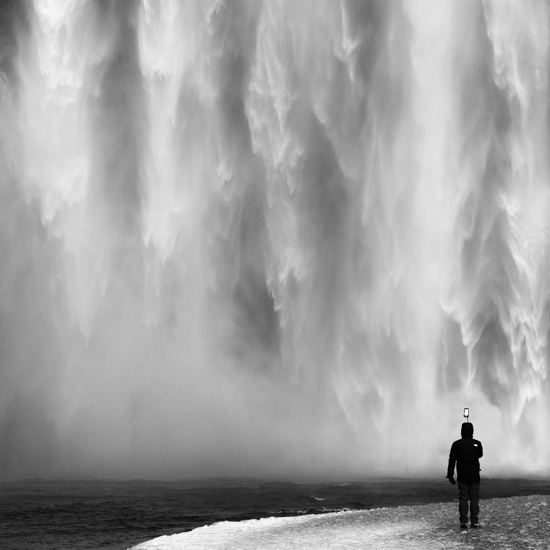 Black-and-white photograph of a tourist holding a phone on a selfie stick before the immense curtain of Skógafoss waterfall in Iceland.