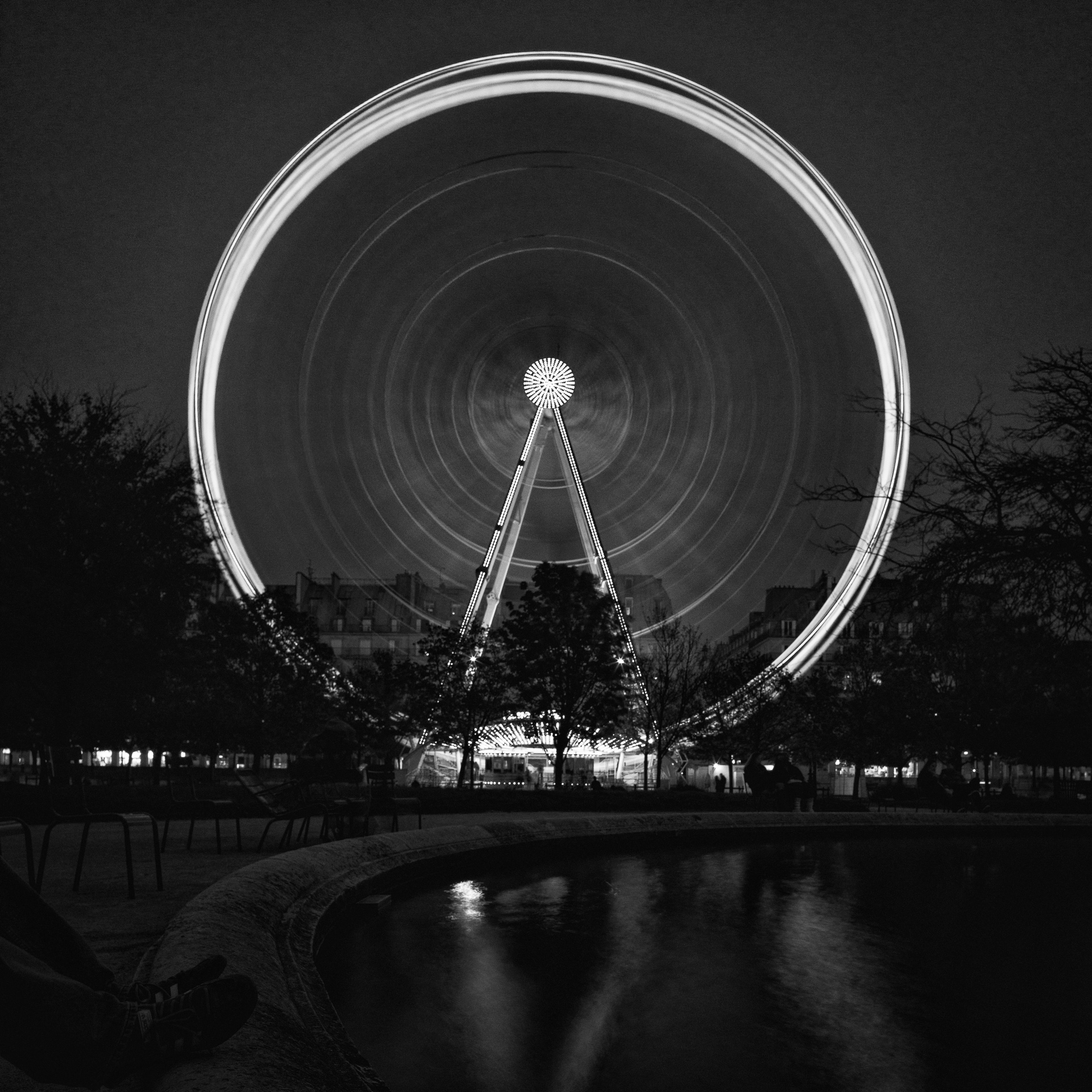 Black-and-white long exposure of the Grande Roue de Paris at night, its illuminated wheel rendered as concentric circles above a dark reflecting pool.