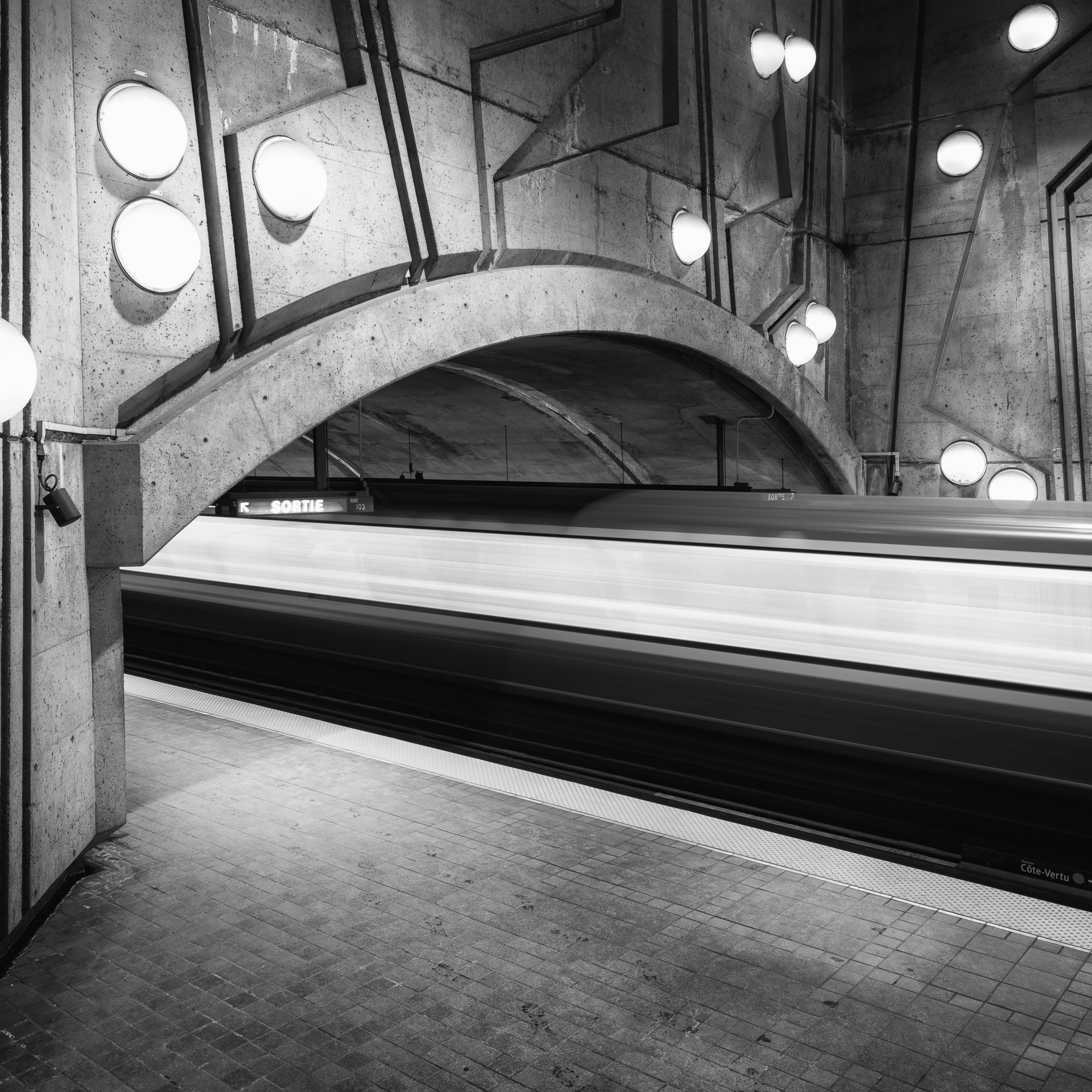 Black-and-white long exposure of a Montreal Metro train passing through De La Savane station, rendered as a horizontal blur beneath the station’s concrete arches and round lights.