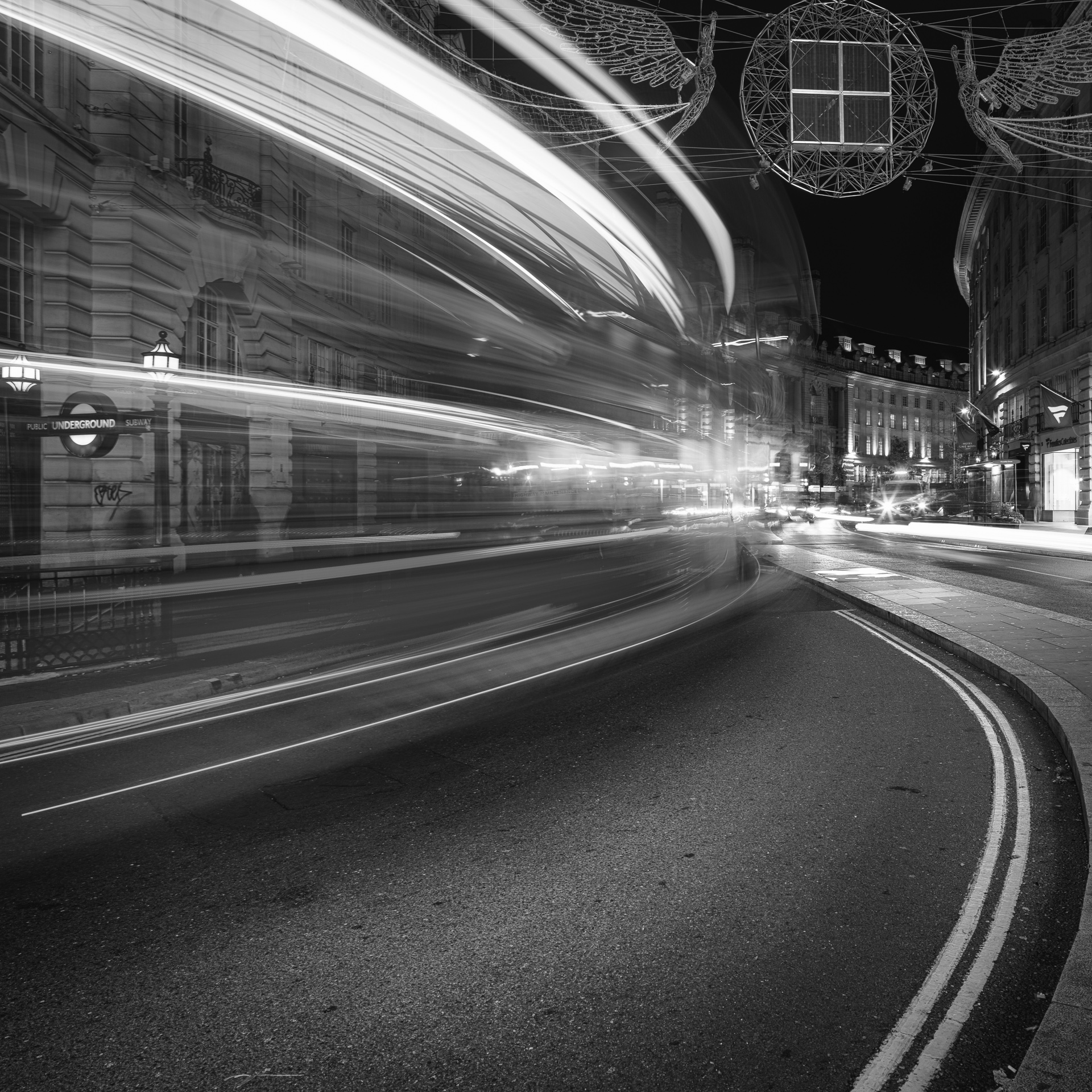 Black-and-white long exposure of a double-decker bus passing along Regent Street at night, leaving bright light trails through the curve of the road.