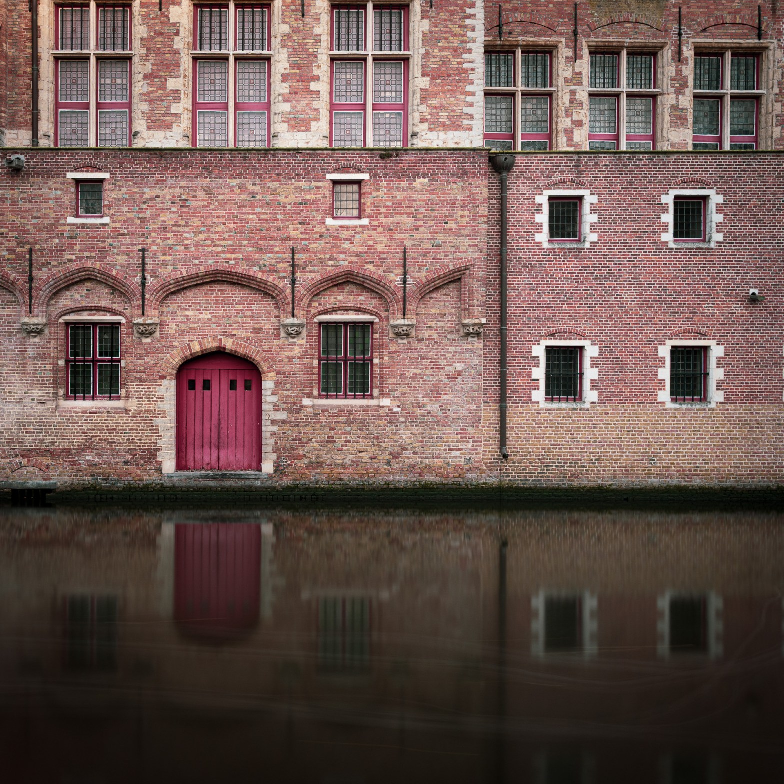 Red door in a historic brick facade reflected in a canal in Bruges, Belgium.