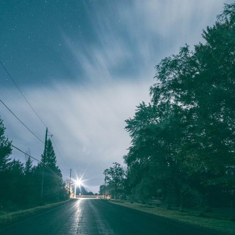 Square crop of a long-exposure night photograph showing a wet road, passing car lights and a lightning-lit storm moving into the distance.