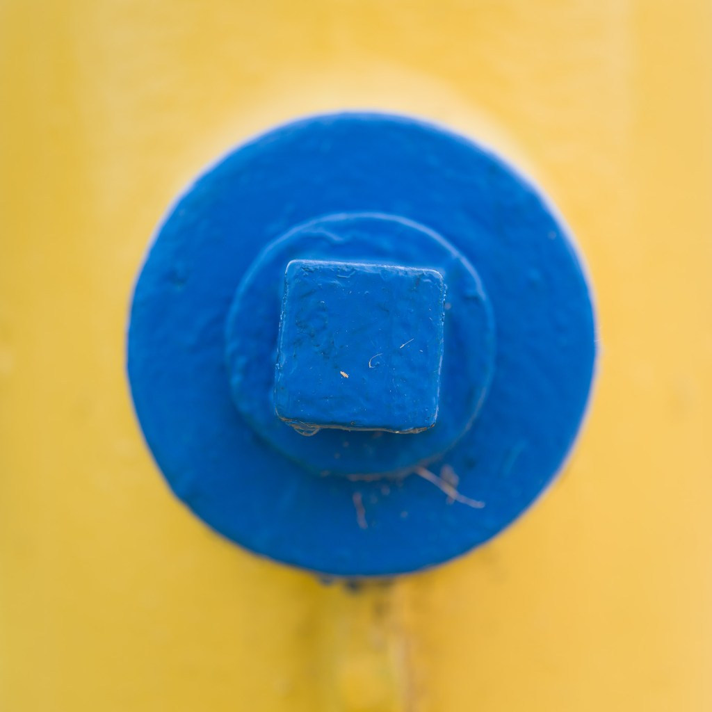 Close-up of a blue-painted circular cap with a square centre on a yellow fire hydrant.