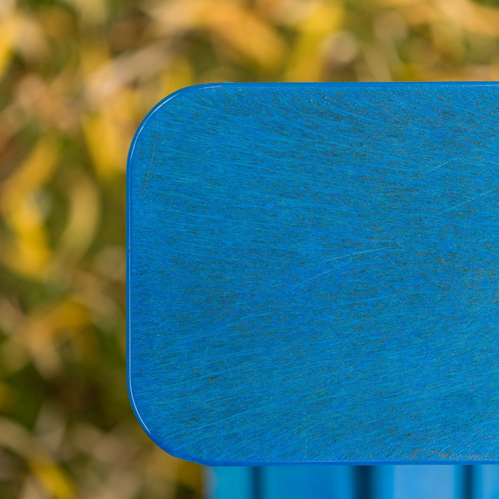 Close-up of a weathered blue Adirondack chair armrest with blurred yellow autumn foliage behind it.