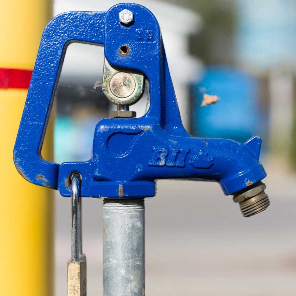 Blue-painted outdoor water spigot mounted on a galvanized post, with a yellow post in the background.