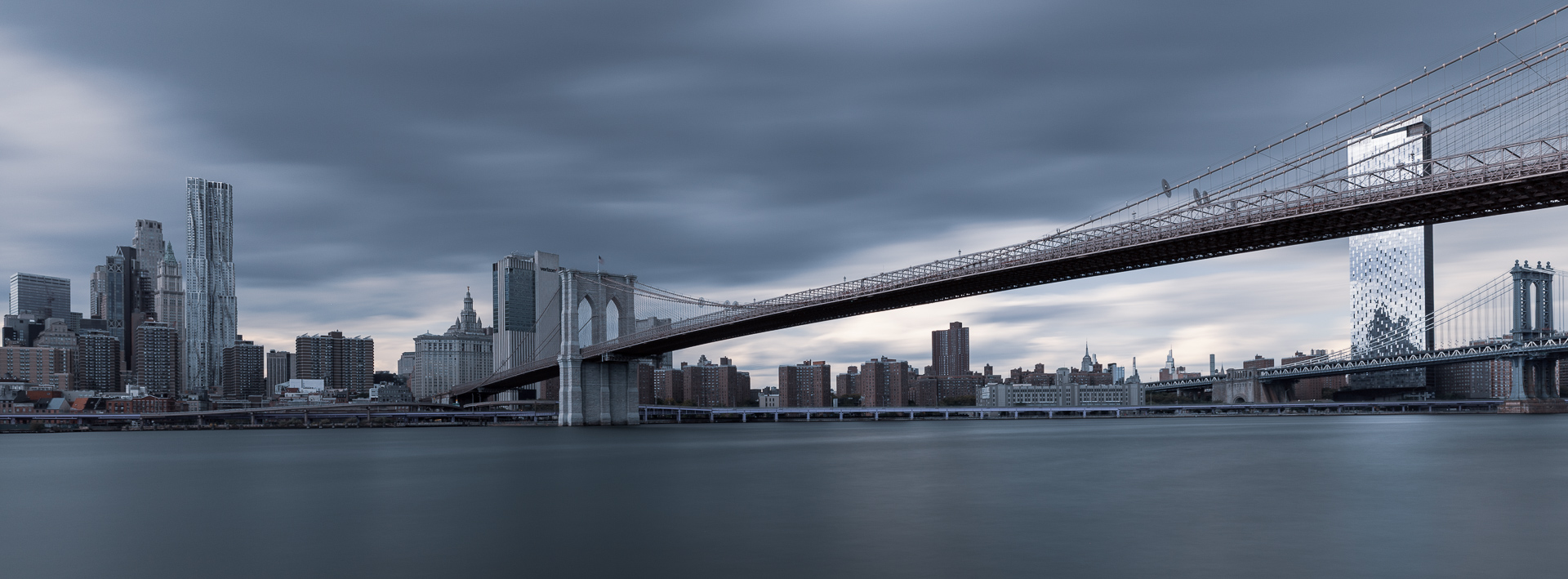 Long-exposure view of the Brooklyn Bridge spanning the East River toward Lower Manhattan, with the Manhattan Bridge visible at right beneath streaked clouds.