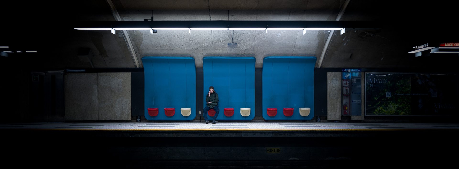 Lone passenger seated beneath bright blue sculptural panels on the platform at Plamondon station in the Montréal Metro.