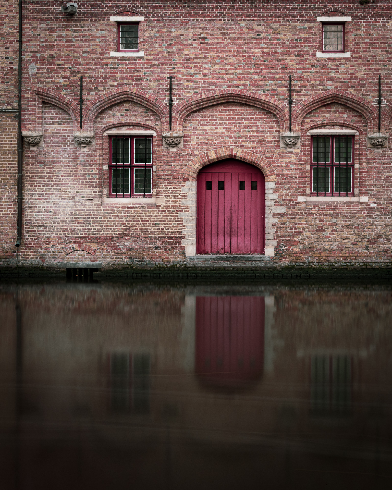 Red doorway in a historic brick canal building in Bruges, reflected in the dark water below.