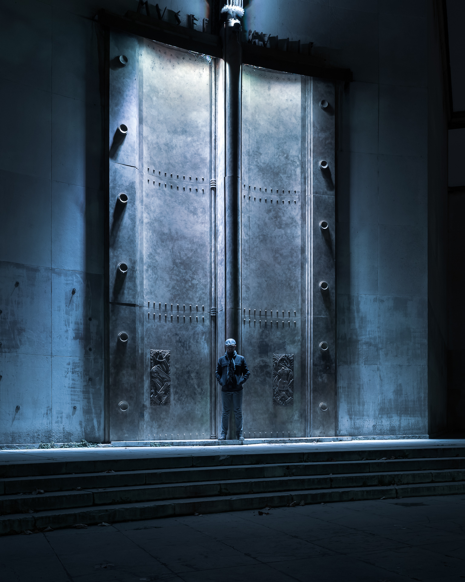 Night self-portrait of a solitary figure standing beneath illuminated monumental metal doors at the Palais de Tokyo in Paris.