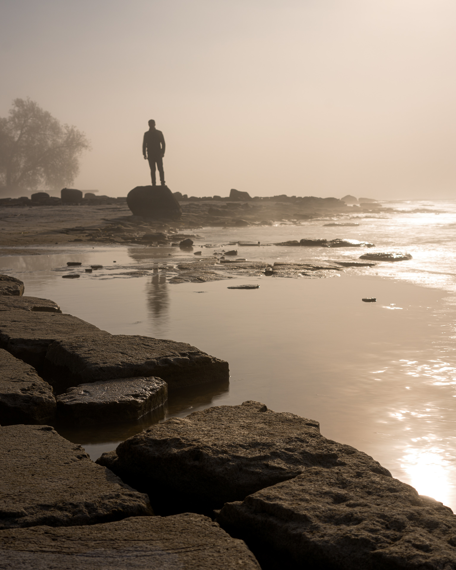 Silhouetted figure standing on a shoreline boulder at sunrise, surrounded by rocks, shallow water and warm mist on Lake Ontario in Kingston.